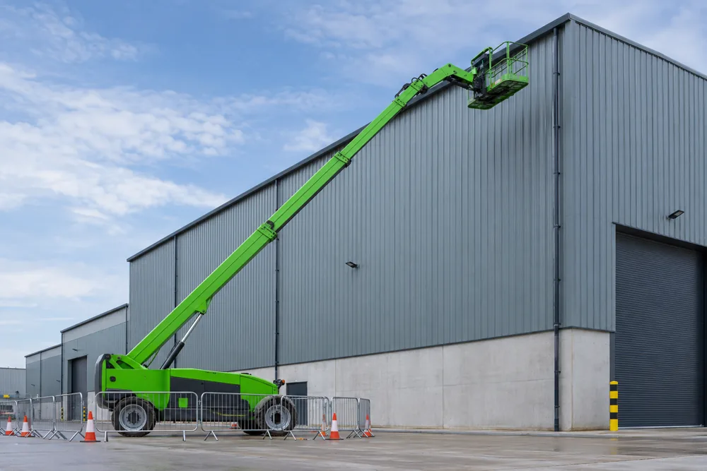 Green telescopic boom lift extended towards a warehouse roof behind safety barriers in Milton Keynes